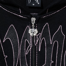 Load image into Gallery viewer, Close up of a black zip hoodie with black embroidered Revenge arch logo with pink contrast stitch outline embroidery and a silver R zipper puller

